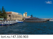 A fragment of the seaport in Yalta. In the center of the frame, an elegant and modern yacht called Orion is moored, painted dark gray with an orange line along the waterline. Nearby are boats and other vessels, and in the background are visible buildings of the port infrastructure, including the control tower with a characteristic dome. The sea is bright and rich blue, light ripples on the water indicate a fresh breeze. On the concrete wall behind is the inscription WELCOME, which emphasizes the tourist character of the place.The photo conveys the atmosphere of active marine life and the modern Yalta coast. (2021 год). Редакционное фото, фотограф Анна Кривицкая / Фотобанк Лори