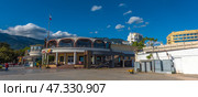 A fragment of the Yalta embankment, one of the most popular resort towns in Crimea. In the foreground is a modern building with a round façade and stained-glass windows, where cafes and restaurants are located. On the left is part of the pedestrian zone with passers-by, vacationers and strollers along the shore. In the background, the green mountains of the Crimean ridge rise, and the sky is decorated with living clouds and a flying bird. The Russian flag on the mast emphasizes the formality and maritime character of the place.The decor conveys an atmosphere of relaxation, resort activity and harmony with the natural landscape. (2021 год). Редакционное фото, фотограф Анна Кривицкая / Фотобанк Лори