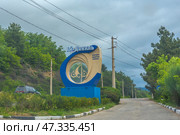 Entrance sign of the resort complex "Ai-Danil", located in Crimea. The sign is made in a modern style: a large decorative structure in the form of a ring, made in beige and blue tones, inside which is the silhouette of a sailboat - a symbol of sea recreation and travel. The inscription reads: "Ai-Danil - a health resort complex". The sign is installed against the background of a green wooded slope, along the road going towards the mountains. In the frame - a cloudy sky and power lines, emphasizing the remoteness and naturalness of the area. Стоковое фото, фотограф Анна Кривицкая / Фотобанк Лори
