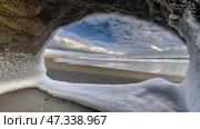 Icy coastal cave view: winter beach scene with clouds and ocean waves. Стоковое AI-изображение, AI-художник Olga Didraga / Фотобанк Лори