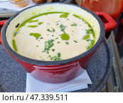 A portion of creamy soup served in a small red saucepan on a grey plate. The soup has a delicate, creamy consistency, topped with drops of aromatic oil (perhaps basil or olive) and finely chopped fresh herbs. All this creates an appetizing and elegant appearance. This dish is usually served in cafes or restaurants specializing in modern European cuisine. Стоковое фото, фотограф Анна Кривицкая / Фотобанк Лори