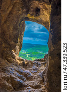 A view from inside the rock cave of the Chelter-Marmara complex onto the vast expanses of Crimea. The uneven stone window seems to frame the landscape: green hills, forests and valleys stretch into the distance, and the sky is covered with dramatic clouds. The contrast between the harsh darkness of the stone and the rich natural palette outside the window creates the effect of deep silence and concentration. A similar view could be observed by the monks who lived here in solitude and prayer. Стоковое фото, фотограф Анна Кривицкая / Фотобанк Лори