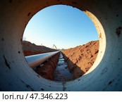 A large pipeline is under construction, visible through a circular concrete culvert. The surrounding area is an open excavation site with earthworks, illuminated by morning sunlight. Стоковое AI-изображение, AI-художник Sergey Grishin / Фотобанк Лори