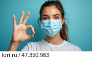 A young woman demonstrates an okay gesture while wearing a light blue mask. The bright blue background highlights her expression of positivity and adherence to health guidelines during the pandemic. Стоковое AI-изображение, AI-художник Sergey Grishin / Фотобанк Лори