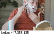 Senior Man shaving his face. Old male shaves beard and mustache with shaver. Elderly gray haired man looking at the mirror, applies shaving cream foam and using razor. Skin hygiene. Daily body care. Стоковое видео, видеограф Марина Демидюк / Фотобанк Лори