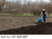 An elderly farmer cultivates soil in early spring, runs a walk-behind tractor machine for ploughing the ground beds on spring day in the backyard on a farm. Стоковое фото, фотограф Ирина Михайличенко / Фотобанк Лори