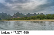 On the bank of a calm river, there is green vegetation - trees, bushes. Picturesque mountains against the sky and clouds. Ripples on the shiny surface of calm water. China. Li River. Li Jiang. Guilin. Стоковое фото, фотограф Вера Ивановна Кубарева / Фотобанк Лори