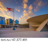 Doha, Qatar - Jan 06 2024, a view of flagpole with the flag of Qatar, fluttering in the wind, with the National Museum of Qatar on background, at evening with cloudy sky, Doha, Qatar. Редакционное фото, фотограф Сергей Богомяко / Фотобанк Лори