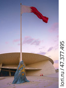 Doha, Qatar - Jan 06 2024, a view of flagpole with the flag of Qatar, fluttering in the wind, with the National Museum of Qatar on background, at evening with cloudy sky, Doha, Qatar. Редакционное фото, фотограф Сергей Богомяко / Фотобанк Лори