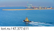 Manama, Bahrain - Jan 08 2024, Panoramic view of passing tugboat, a boat, in the background a commercial international port with cranes, pier, buildings and containers, in the daytime, Manama, Bahrain. Стоковое фото, фотограф Сергей Богомяко / Фотобанк Лори