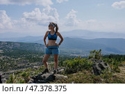 Tourist Woman in blue shorts and sports bra stands on a stone against the backdrop of summer landscape in the mountains. Стоковое фото, фотограф Екатерина Якименко / Фотобанк Лори