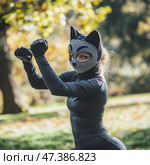 A man in a tight black suit with a hood in the form of a cat mask. The mask has ears and a pattern imitating a cat's face, leaving only the eyes open. The character has taken a fighting stance, with his hands raised, as if ready to attack or defend. The scene takes place outdoors, in a park or forest, with autumn trees and soft natural light. The image is reminiscent of a superhero or a cosplay character. Стоковое AI-изображение, AI-художник Анна Кривицкая / Фотобанк Лори