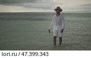 A young man in light white clothes and a hat walks barefoot in the rain on the water of the sea. Stormy evening clouds are in the background. Maldives. Стоковое видео, видеограф Aleksey Kidisyuk / Фотобанк Лори