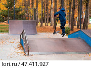 A teenager on a stunt scooter on a playground in a skate park. A child in a tracksuit performs a trick in an autumn park while jumping. Стоковое фото, фотограф Photojuli / Фотобанк Лори