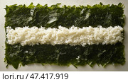 Glossy nori sheet with sushi rice and floral accents.Глянцевый лист нори уложен ровным слоем, а сверху равномерно распределен тонкий, нежный слой риса для суши. Свежие цветочные акценты дополняют элегантную подачу, а рядом стоит чашка чая. Стоковое AI-изображение, AI-художник Katsiaryna Repich / Фотобанк Лори