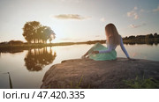 A lonely girl sits on a lake on a stone on a sunset background. Video on the move. Стоковое видео, видеограф Mikhail Davidovich / Фотобанк Лори