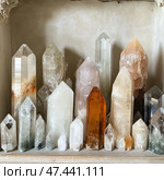 A collection of natural crystals of various shapes, sizes and colours, neatly arranged on a shelf against the wall. The crystals are pointed, hexagonal and range in colour from clear and white to soft pink, amber, smoky and pale green. The lighting is soft and natural, highlighting the texture, purity and internal inclusions in the minerals. The composition looks aesthetically pleasing, harmonious and can be associated with energy, healing, spiritual practices or simply collecting natural beauty. Стоковое AI-изображение, AI-художник Анна Кривицкая / Фотобанк Лори
