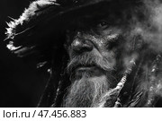 Expressive black and white portrait of an elderly man in an ethnic or shamanic image. His face is covered with textured paint or dirt, his beard is thick and gray, and his hair is decorated with braided elements, beads and feathers. The feathered headdress enhances the mystical and ritualistic mood. The man's gaze is penetrating, serious, as if carrying ancient wisdom. The composition and lighting create the effect of fog or smoke, enhancing the feeling of mystery and antiquity. Overall, the image resembles the archetype of a shaman, warrior or old chieftain. Стоковое AI-изображение, AI-художник Анна Кривицкая / Фотобанк Лори