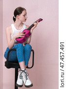 Young female musician plucking ukulele, sitting on bar stool, looking at strings musical instrument. Стоковое фото, фотограф А. А. Пирагис / Фотобанк Лори