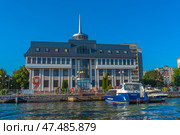 The building of the river terminal in Kaliningrad, view from the water. The architecture is modern, with a symmetrical façade, a high spire and panoramic windows. Boats are moored in front of the building, including a white and blue passenger boat with the inscription "Rosmorport" and the name "Victoria". The clear sky, reflections on the water and bright lighting give the frame a summery, sunny mood. On the facade of the building you can see a banner with the number "9" - a reference to May 9 (Victory Day). This is a landmark place for river cruises along the Pregolya. (2020 год). Редакционное фото, фотограф Анна Кривицкая / Фотобанк Лори