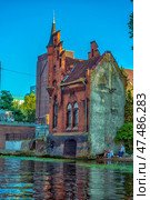 A picturesque red brick Gothic building standing right on the river bank in Kaliningrad. The corner turret with a high spire and decorative battlements give the building the appearance of a small castle or watchtower. The building can be recognized as one of the old water towers or security structures typical of Königsberg architecture. In the foreground is a water surface with a reflection of the building, and on the right are people relaxing with a dog. In the background are modern and historical brick houses. (2020 год). Редакционное фото, фотограф Анна Кривицкая / Фотобанк Лори