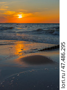 A picturesque sunset over the Baltic Sea. The sun slowly sinks below the horizon, painting the sky and water in rich shades of orange, yellow and blue. The waves gently roll onto the sandy shore, reflecting the golden light. In the foreground are wet sand covered with surf foam and wooden riots extending into the water. The atmosphere of calm, solitude and natural harmony makes the scene ideal for illustrating themes: relaxation, nature, the Baltic, meditation, travel. Стоковое фото, фотограф Анна Кривицкая / Фотобанк Лори