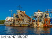 An old industrial transport vessel moored off the coast of Kaliningrad. This is a dredger called "Alexander Uvarov", according to the inscription on the side. The vessel has a massive structure with signs of rust and wear, which gives it an industrial character. It is equipped with cargo and working mechanisms: a conveyor system, manipulators, ladders. In the background is a blue industrial hangar and a mast with the Russian flag. The bright sunny day emphasizes the texture of the metal, water and rust. (2020 год). Редакционное фото, фотограф Анна Кривицкая / Фотобанк Лори