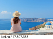 Островной пейзажю Женщина в шляпе сидит на фоне моря и бело-голубых домов острова Санторини. Стоковое AI-изображение, AI-художник Башкирова Анна / Фотобанк Лори