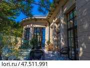 A cozy outdoor terrace of a modern building, surrounded by a glass fence and pine trees. The architecture is neoclassical: light stone, tall windows in black frames, columns with ribbed details. The terrace is furnished with designer rattan and black plastic chairs and tables. Light penetrates through the tree crowns, casting beautiful shadows on the walls and floor. The space looks like part of a luxury hotel, restaurant or villa — perfect for relaxing and connecting with nature. Стоковое фото, фотограф Анна Кривицкая / Фотобанк Лори