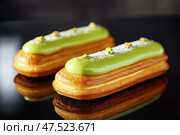 Pistachio-glazed eclairs rest symmetrically on a gleaming black surface. Their glossy, vibrant green coating contrasts dramatically with the sleek, reflective background. Стоковое AI-изображение, AI-художник Nart / Фотобанк Лори