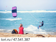 Kiteboarder riding kite across the waves of Flag beach on vacations on Fuerteventura, Canary islands while her girlfriend watches him from the sandy beach, sunbathing. Стоковое фото, фотограф Matej Kastelic / Фотобанк Лори