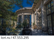 A cozy outdoor terrace of a modern building, surrounded by a glass fence and pine trees. The architecture is neoclassical: light stone, tall windows in black frames, columns with ribbed details. The terrace is furnished with designer rattan and black plastic chairs and tables. Light penetrates through the tree crowns, casting beautiful shadows on the walls and floor. The space looks like part of a luxury hotel, restaurant or villa — perfect for relaxing and connecting with nature. Стоковое фото, фотограф Анна Кривицкая / Фотобанк Лори