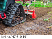 A tractor equipped with a red rotary cultivator actively prepares the soil in a rural field. The equipment loosens the earth, preparing it for planting and cultivation. Стоковое фото, фотограф Коханова Наталья / Фотобанк Лори