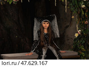 A child girl model showcases a stunning dark costume while seated against a backdrop of natural elements. Her long hair flows gracefully, complementing the enchanting atmosphere of the moment. Стоковое фото, фотограф Aleksandr Golubev / Фотобанк Лори