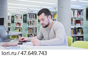 Focused guy student preparing for an exam on a laptop in the university library, looking for sources of information in textbooks. Стоковое видео, видеограф Яков Филимонов / Фотобанк Лори