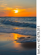 A picturesque sunset over the Baltic Sea. The sun slowly sinks below the horizon, painting the sky and water in rich shades of orange, yellow and blue. The waves gently roll onto the sandy shore, reflecting the golden light. In the foreground are wet sand covered with surf foam and wooden riots extending into the water. The atmosphere of calm, solitude and natural harmony makes the scene ideal for illustrating themes: relaxation, nature, the Baltic, meditation, travel. Стоковое фото, фотограф Анна Кривицкая / Фотобанк Лори