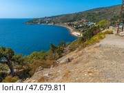 A magnificent view of the Crimean coast. From above, a panorama of the curved coastline, covered with sparse vegetation, rocks and junipers, opens up. In the distance, a small town or village with white houses spread out along the slope of a hill is visible. The Black Sea is deep blue, calm, with slight glare from the sun. On the horizon, closer to the center of the frame, an old sea platform or pier is noticeable. This place conveys an atmosphere of privacy, tranquility and southern warmth. Стоковое фото, фотограф Анна Кривицкая / Фотобанк Лори