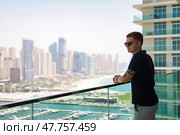 A guy stands on a balcony, observing the stunning Dubai skyline filled with skyscrapers and the waterfront. He wears sunglasses and has a relaxed demeanor, enjoying the vibrant atmosphere. Стоковое фото, фотограф Aleksandr Golubev / Фотобанк Лори