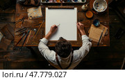An overhead view captures an artist holding a pencil over a blank canvas, surrounded by paintbrushes, paints, and art supplies on a wooden table. Стоковое AI-изображение, AI-художник Данила Большаков / Фотобанк Лори