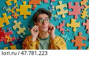 Young man wearing glasses and a yellow jacket stands in front of a large puzzle wall, studying the pieces intently. Стоковое AI-изображение, AI-художник Данила Большаков / Фотобанк Лори