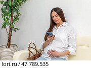 Pregnant woman using a smartphone and gently touching her belly while relaxing on couch in hospital. Happy young expectant mother. Family planning and IVF centers concept. High quality. Стоковое фото, фотограф Ирина Михайличенко / Фотобанк Лори