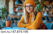A girl with glasses in a vibrant beanie smiles contentedly while surrounded by books and art tools in a cozy cafe setting. Стоковое AI-изображение, AI-художник Данила Большаков / Фотобанк Лори