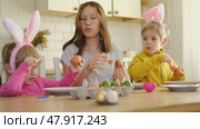 Children siblings painting Easter egg sitting together at table. Easter Family traditions. Teen girl teaching happy little brother and sister to dye, decorate eggs with paints. Preparation to holidays. Стоковое видео, видеограф Марина Демидюк / Фотобанк Лори