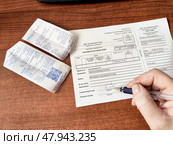 Kirov, Russia - March 12, 2025: Individual completes a transaction form at a wooden desk with receipts in hand. Редакционное фото, фотограф Кривошеина Елена Леонидовна / Фотобанк Лори
