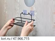 Hands are installing black metal bathroom shelf onto wall mounted transparent adhesive hooks by aligning hooks with designated attachment points. Стоковое фото, фотограф Владимир Григорьев / Фотобанк Лори
