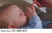 A woman's hand feeds porridge from a spoon baby sitting in a highchair. The baby's face is covered with food. Useful food. Breakfast porridge. Стоковое видео, видеограф Воловодов Владимир / Фотобанк Лори