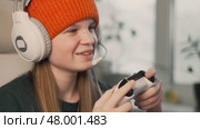 Girl enjoying video game in headphones with microphone, sitting comfortably at desk. Bright daylight, stylish tech gadgets and vibrant beanie create positive modern vibe. Стоковое видео, видеограф Енваев Андрей / Фотобанк Лори
