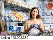 Young woman choosing decorative plate with colorful pattern in souvenir shop. Стоковое фото, фотограф Яков Филимонов / Фотобанк Лори