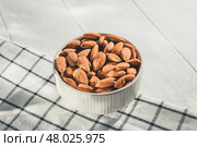 Shelled almonds are piled high in a small white bowl resting on a checkered cloth, creating a simple yet appealing image of healthy snacking. Стоковое фото, фотограф Ольга Ильина / Фотобанк Лори