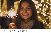 Smiling Girl with Fire Sparkler Glow in the Winter Season. pleased happy smiling young 20s woman is holding a bright sparkler in a festive winter setting. Glowing blur bokeh light on background. Стоковое AI-видео, аниматор Марина Демидюк / Фотобанк Лори